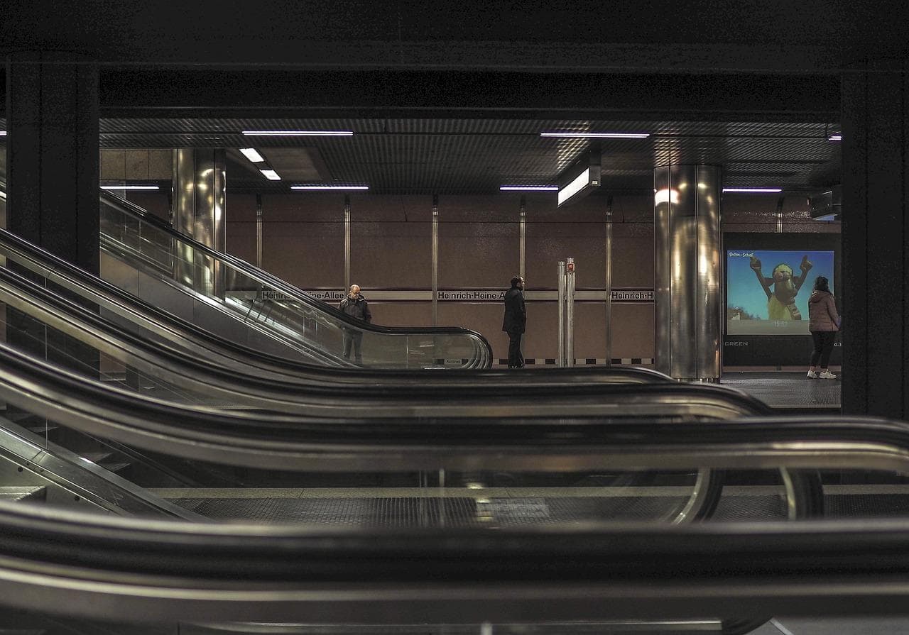 A dimly lit subway station with multiple escalators in the foreground. Three people are standing on the platform, and a large screen displays an image of a person wearing a white robe with arms raised.