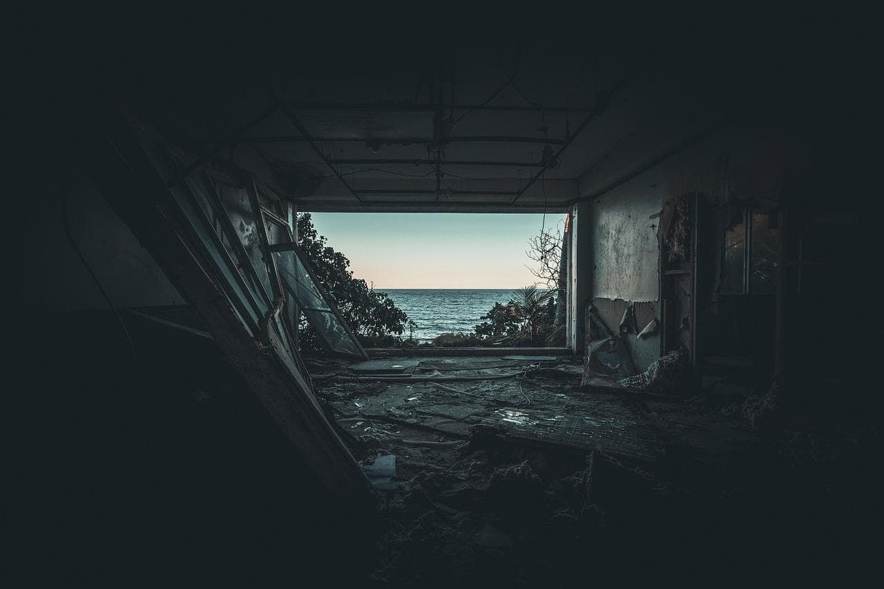 A view of the ocean seen through the broken walls and debris of an abandoned, dilapidated building, with dim light illuminating the ruins.