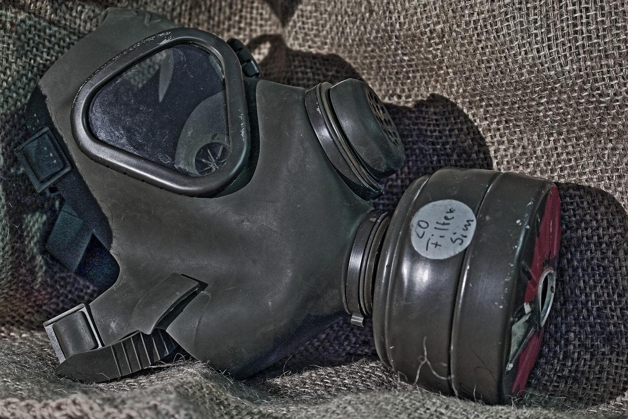 A black gas mask with a filter labeled CO filter Sim rests on rough, brown burlap fabric, reflecting light on its visor.