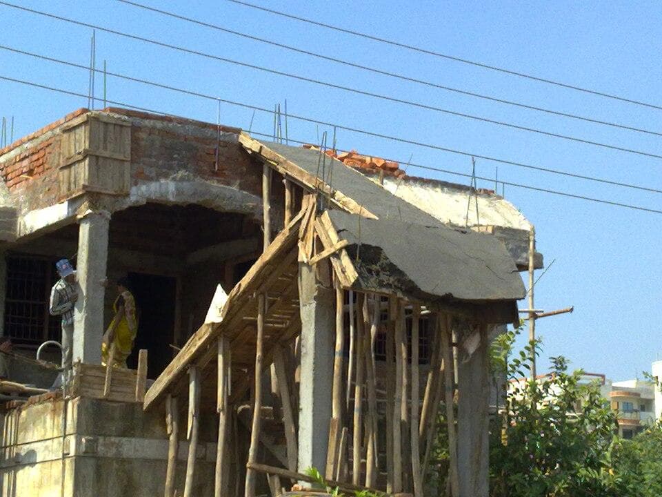 A building under construction with brick walls, exposed concrete pillars, and wooden scaffolding. Two workers are visible on the site, and power lines run above with a clear blue sky in the background.