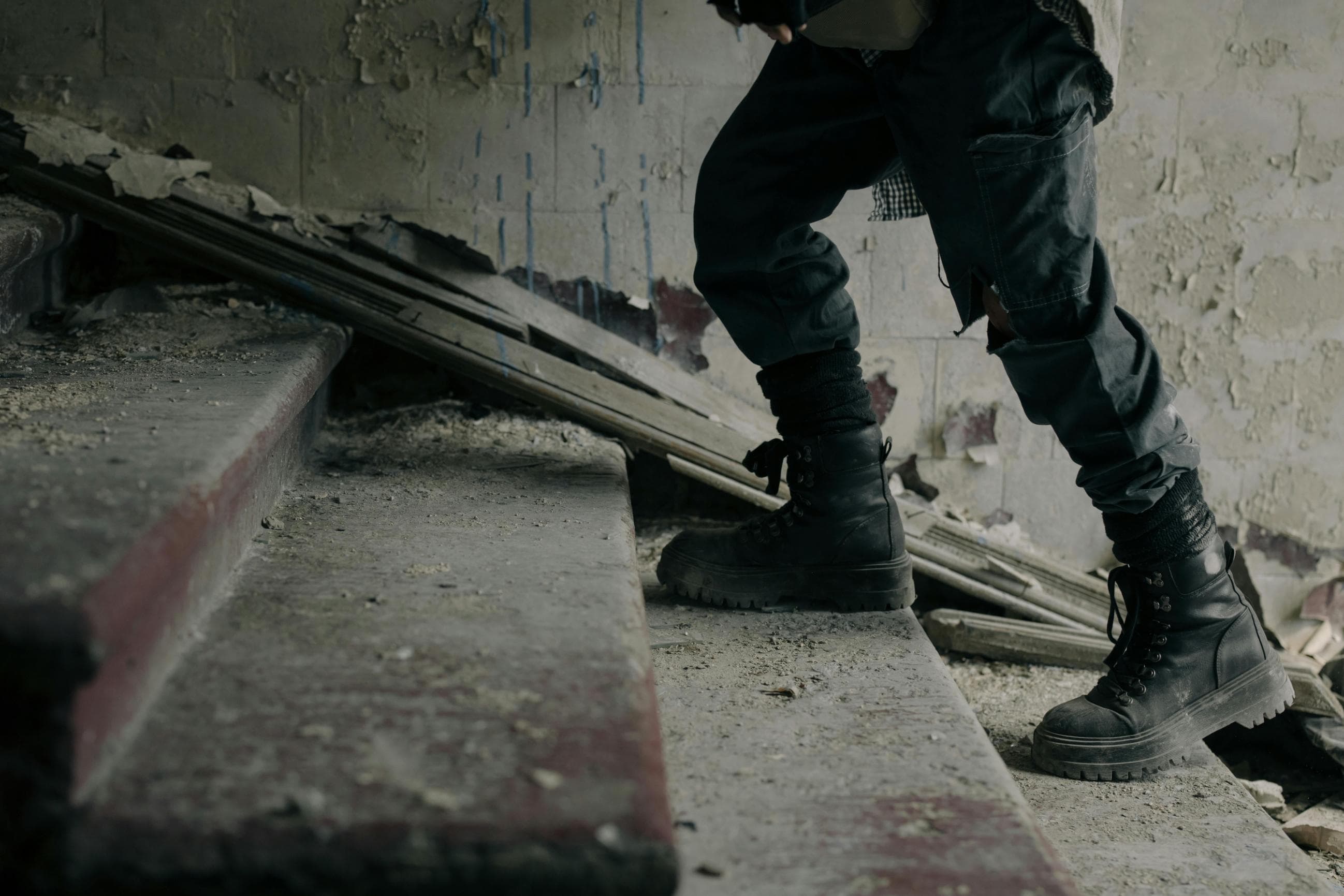 A person wearing black combat boots and dark pants walks up dusty, worn concrete stairs in a dilapidated building with peeling paint and debris scattered around.