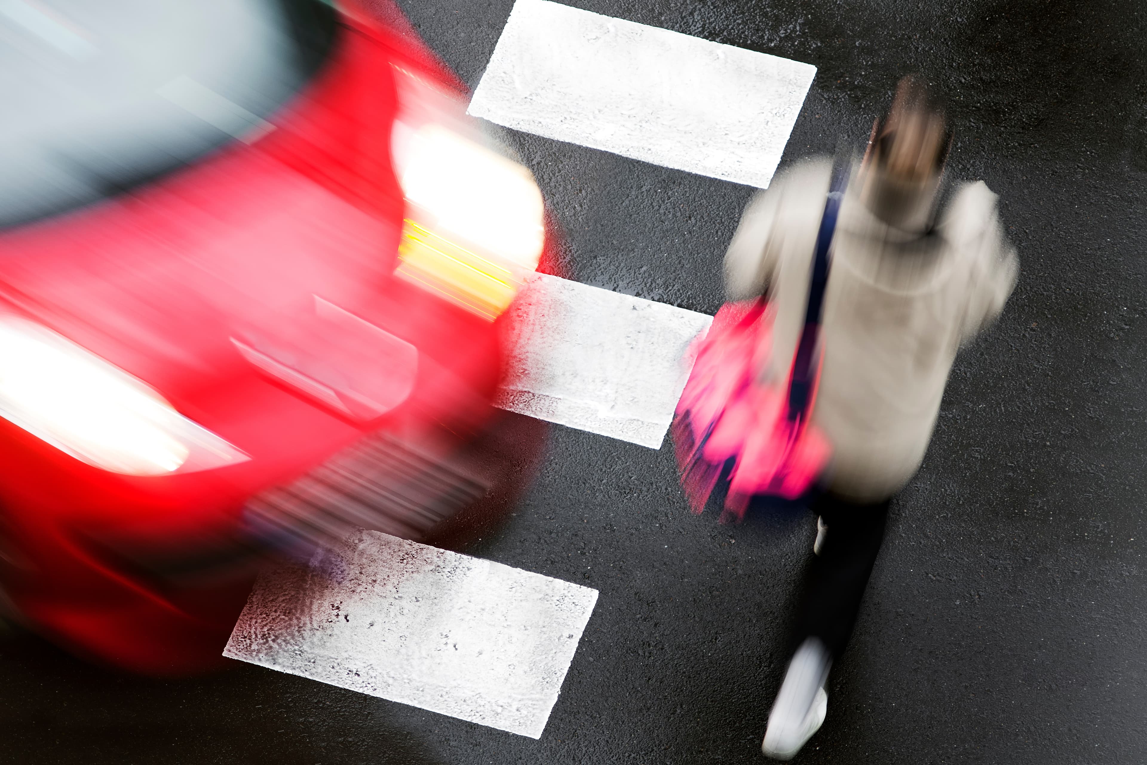 Blurred red car passing close to a pedestrian walking across a crosswalk on a wet street