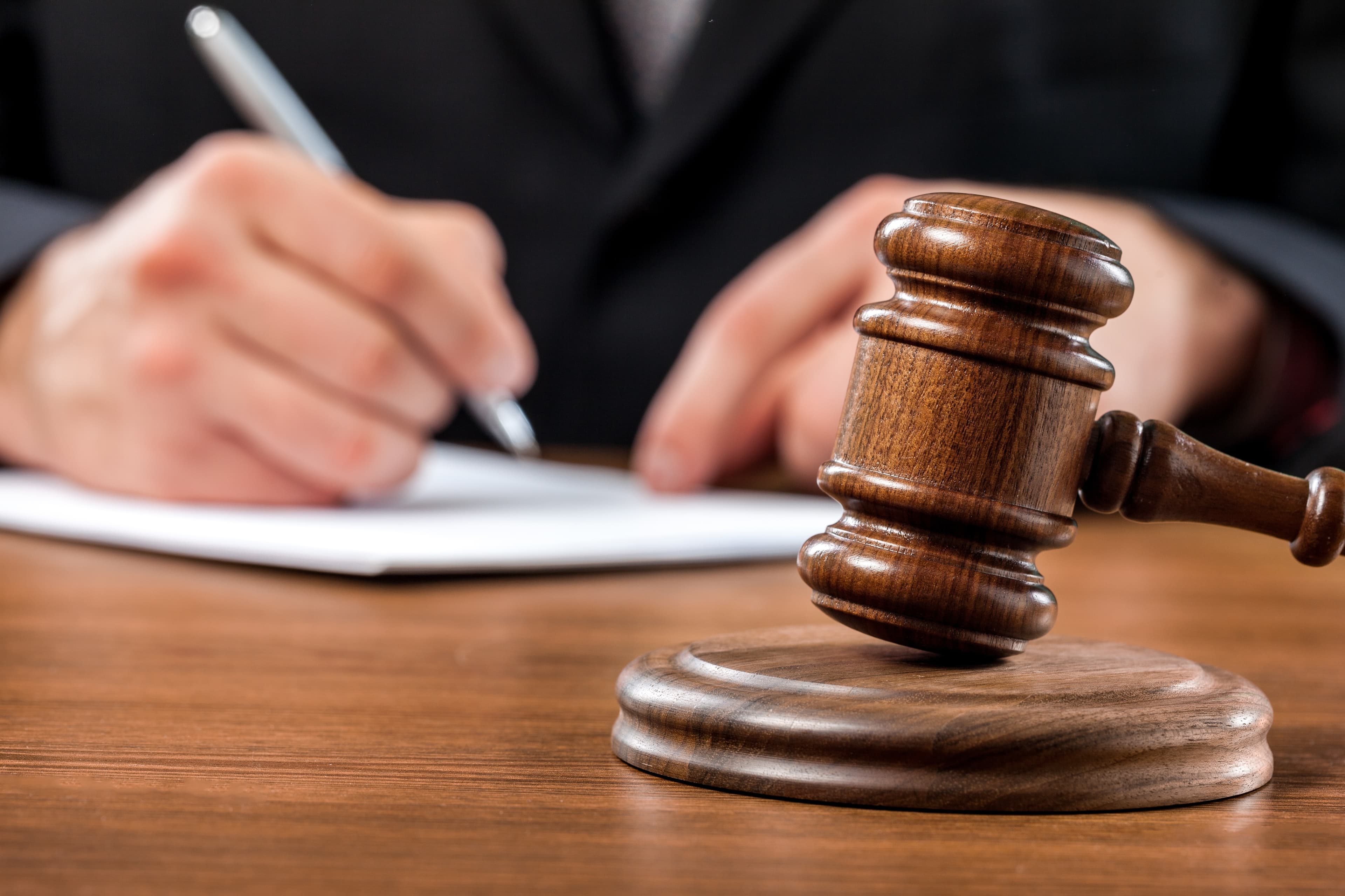 Dark wood gavel resting on a table in the foreground. Lawyer holding a pen over a piece of paper on the same table in the background | Personal Injury | The Sullivan Law Firm