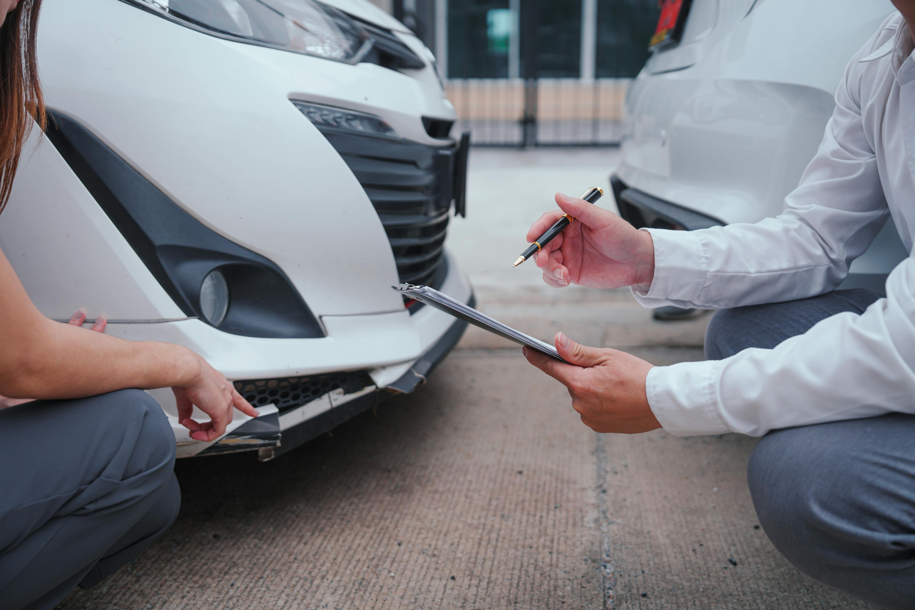 Woman pointing to accident damage on her car, next to a man in business clothes holding a clipboard and taking notes of the accident | Automobile Accidents | The Sullivan Law Firm